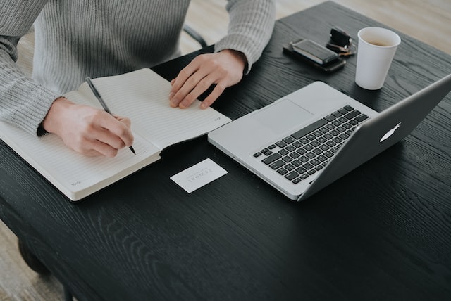 Person writing in a notebook in front of their open laptop